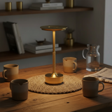 Wooden table with a gold lamp, mugs, and a pitcher in a cozy room setting.