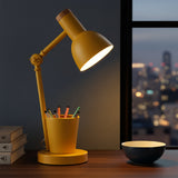 Yellow desk lamp on a table with books and a bowl, city lights visible through a window.